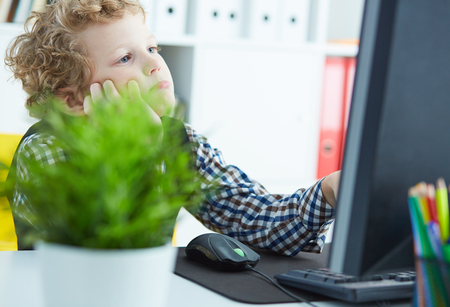 Tired little Caucasian boy propped his head in his hands and looks at the monitor at the officeの写真素材