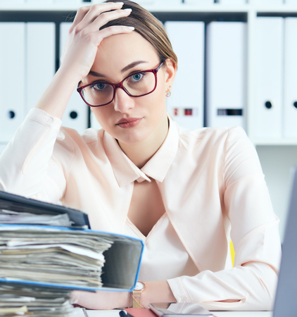 Young tired and exhausted woman in spectacles looks at the camera leaning against mountain of documents propping up her head with her hands.の写真素材