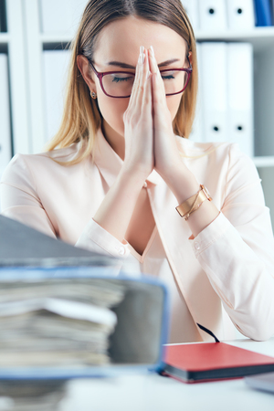 Close up stressed office woman doing praying hand gesture while looking at the pile of documents at her workplace.の写真素材
