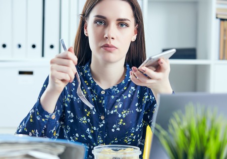 Business woman eating lunch at her workplace looking at the laptop screen. Folders with documents in the foreground. Dedline concept.の写真素材