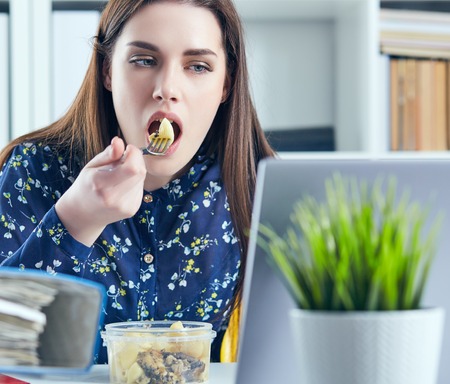 Business woman eating lunch at her workplace looking at the laptop screen. Folders with documents in the foreground. Dedline concept.の写真素材