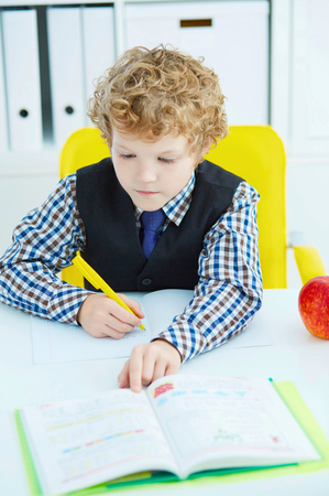 Little Caucasian schoolboy writing in a notebook sitting at a table . Red apple lies next to the table.の写真素材