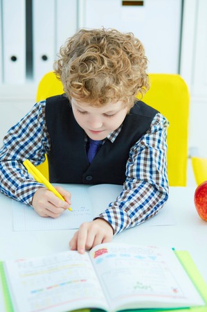 Little Caucasian schoolboy writing in a notebook sitting at a table . Red apple lies next to the table.の写真素材