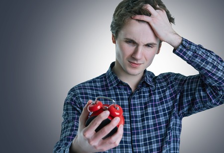 Close-up photo of upset young man with hand in hair holding red alarm clock, isolated over gray background.の写真素材