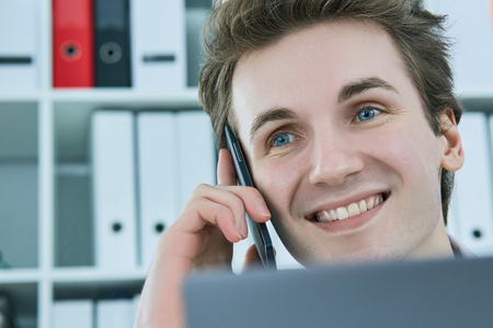 Smiling white collar talking to customer on mobile phone in office at workplace by laptop.の写真素材
