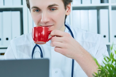 Portrait of friendly male doctor smiling holding a cup of coffee and looking at laptop screen.の写真素材
