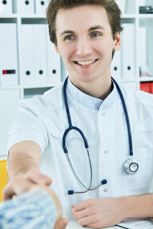 Young male Caucasian doctor shaking hands with patient at doctors bright modern office in hospital.の写真素材
