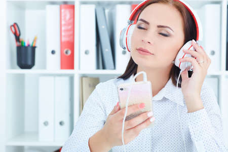 Beautiful Caucasian girl in headphones listening to music from smartphone in office.