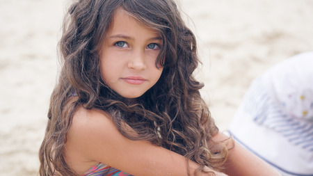 Close-up portrait of a pretty little Hispanic girl with waving in the wind long hair sitting on the beach.の写真素材