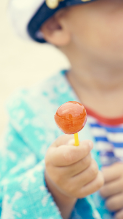 Little boy in a striped t-shirt and captains cap sitting on the beach with candy in mouthの写真素材