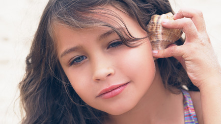 Beautiful little girl with long curly hair listening to sea music in cockleshell on the beach.の写真素材