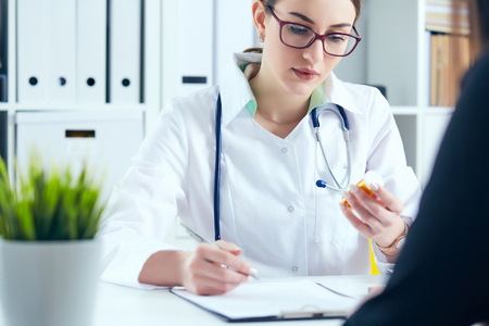Young female medicine doctor in glasses sitting in front of working table holding jar of pills and writing prescription on special form.の写真素材