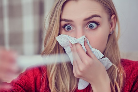 Shocked girl looking on thermometer while sitting on sofa at home.の写真素材