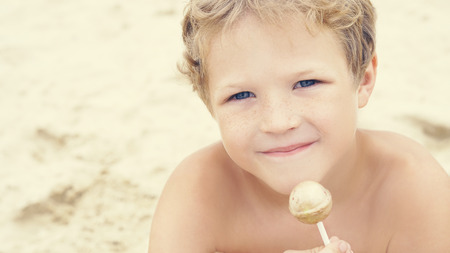 Cute little Caucasian boy at beach with a colorful candy in hand sitting on tropical beach and looking into the camera. Vacation concept.の写真素材