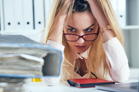 Tired and exhausted woman in spectacles looks at the mountain of documents propping up her head with her hands.の写真素材