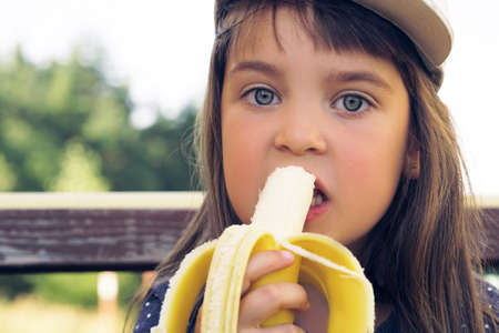 Attractive caucasian child with long hair, in blue t-shirt and white cap eating banana and smiles on a picnic in park.の写真素材