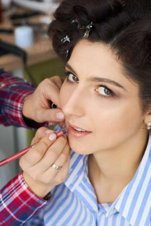 The make-up artist doing a makeup for bride. Makeup artist applies red lipstick . Beautiful woman face. Hand of make-up master, painting lips of young beauty model girl . Make up in processの写真素材