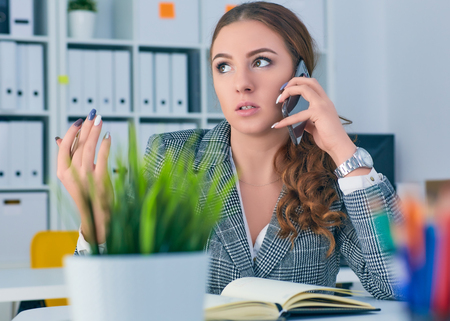 Young serious Caucasian businesswoman sitting at desk in office, speaking on the phone.の写真素材