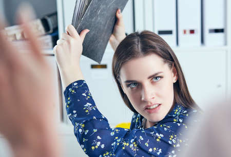 Angry business woman with folder trying to hit his colleague in office. Stressful situations at work.の写真素材