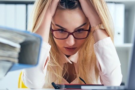 Tired and exhausted woman in spectacles looks at the mountain of documents propping up her head with her hands.の写真素材