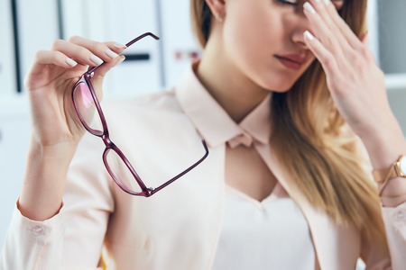 Tired Caucasian businesswoman holding eyeglasses and massaging nose bridge. Girl feeling discomfort from long wearing glasses at workplace.の写真素材