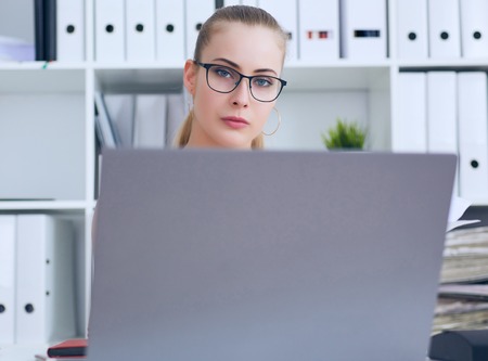 Young beautiful Caucasian businesswoman in glasses locing in camera while working on notebook computer and business document at office.の写真素材