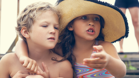 Portrait of children on the beach in summer. Little girl in a hat hugs her brother. They are sitting on the stairs by the sea.の写真素材