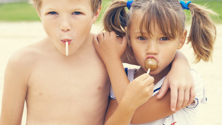 Cute little Latin girl and Caucasian boy necking with a colorful candies in mouths on tropical beach vacationの写真素材