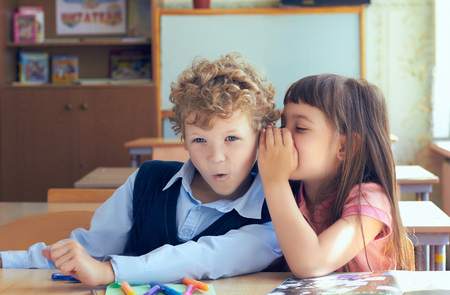 Little girl telling secret to surprised boy in classroom.の写真素材