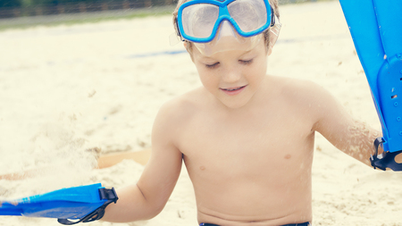 Little curly Caucasian boy with snorkeling equipment sitting on sand on tropical beach.の写真素材