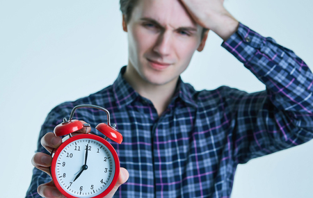Close-up portrait of a troubled young man holding alarm clock isolated over white background.の写真素材