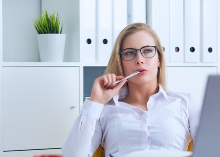 Young Caucasian business woman in glasses looks thoughtfully into the distance holding the pen in her mouth and thinks.の写真素材