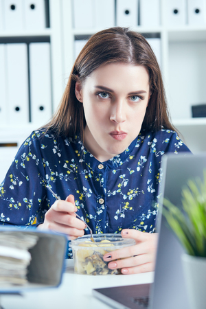 Business woman eating lunch at her workplace looking at the laptop screen. Folders with documents in the foreground. Dedline concept.の写真素材