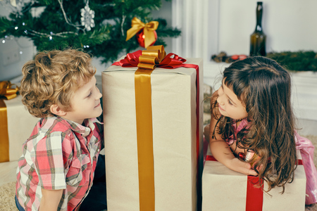 Little girl peeks out from behind big Christmas gift box and looks at smiling boy.の写真素材