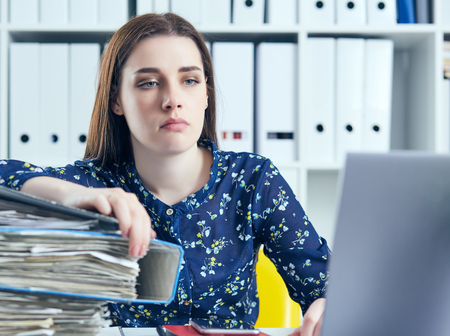Young tired and exhausted woman looks at the camera leaning against mountain of documents propping up her head with her hands.の写真素材