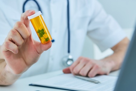 Male medicine doctor hand hold jar of pills and explain how to take pills to patient at worktable. Panacea and life save prescribing treatment legal drug store concept.の写真素材