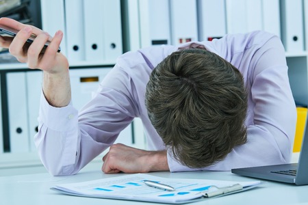 Young businessman lying on the desk in office holding smartphone, keeping face on his hands. Deadline concept. Time to call nine one one.の写真素材