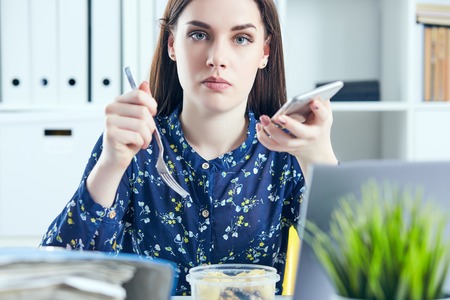 Business woman eating lunch at her workplace looking at the laptop screen. Folders with documents in the foreground. Dedline concept.の写真素材