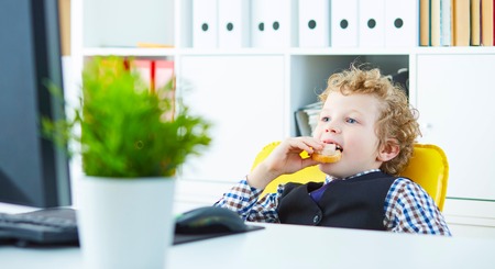 Short break concept at work. Curly Caucasian boy eats a sandwich with sausage in the workplace in the office and looking at the computer screenの写真素材