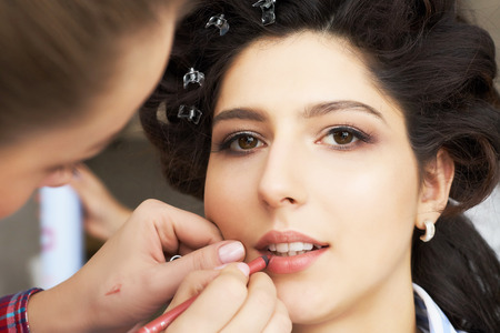 The make-up artist doing a makeup for bride. Makeup artist applies red lipstick . Beautiful woman face. Hand of make-up master, painting lips of young beauty model girl . Make up in processの写真素材