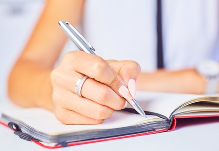 Young woman working at her desk taking notes closeup. Focus on hand writing on a notepad.の写真素材