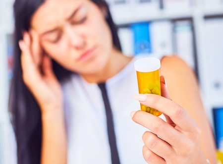 Young sick tired sad woman sitting in workplace holding bottle with pills. Female feeling bad at work.の写真素材