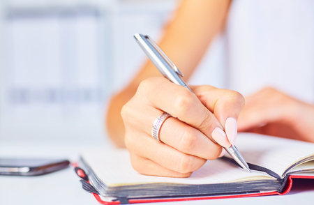 Young woman working at her desk taking notes closeup. Focus on hand writing on a notepad.の写真素材