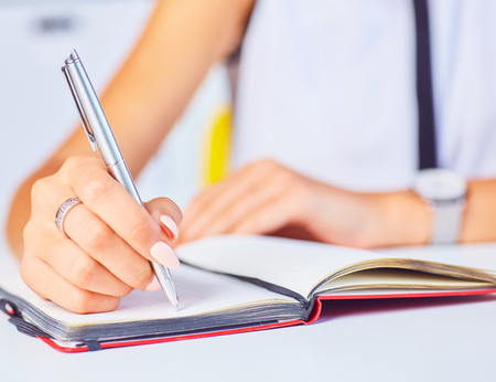 Young woman working at her desk taking notes closeup. Focus on hand writing on a notepad.の写真素材
