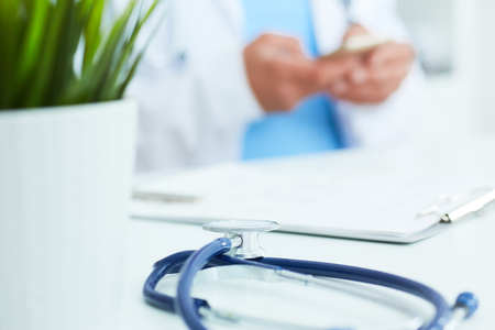 Blue stethoscope lies on the table in the foreground and male doctor hands texting on a smart phone in background.の写真素材
