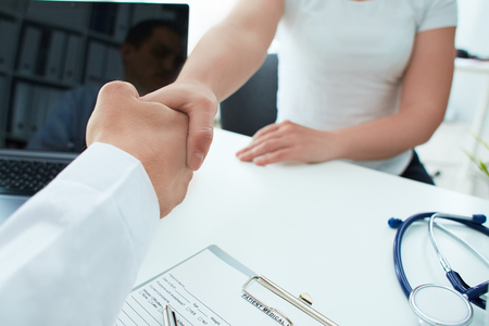 Male doctor take care to female patient and congratulations for his good health. Doctor and patient shaking hands in the office, they are sitting at desk, hands close up.の写真素材