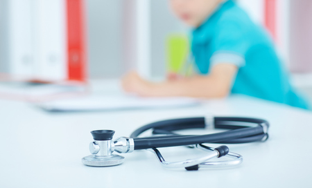 Little boy writes on clipboard sitting at doctors desk. Stethoscope in the foreground. Focus on the stethoscope.の写真素材