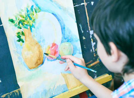 Cute, serious and focused, seven years old boy in blue shirt drawing on canvas standing on the easel. Concept of early childhood education, painting, talent, happy family or parenting.の写真素材