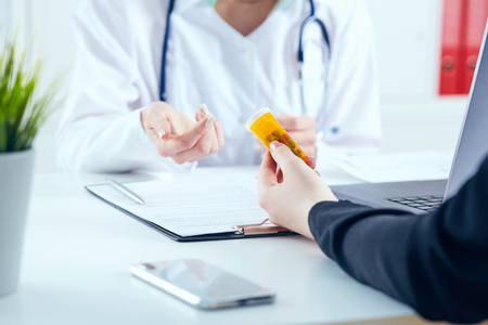 Young female medicine doctor sitting in front of working table holding jar of pills and explains to the patient how to take medication. Medical and pharmacy conceptの写真素材
