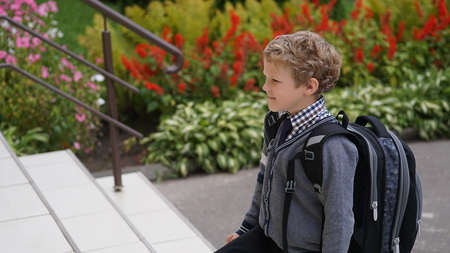 Little Caucasian curly boy in school uniform with backpack comes up on stairs.
